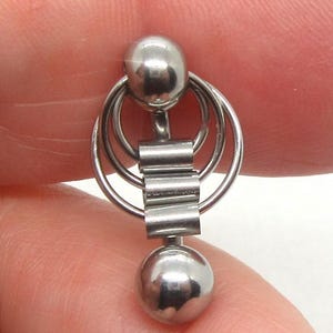 Könnte beinhalten: Ein silberfarbener Körperpiercingschmuck. Der Schmuck besteht aus zwei kugelförmigen Kugeln, die durch Ringe und einen rechteckigen Stab verbunden sind. Das Schmuckstück wird zwischen Fingern gehalten.