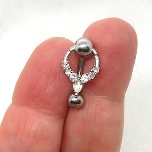 Könnte beinhalten: Ein silberfarbener Bauchnabelring mit einem kreisförmigen Design, das mit kleinen, klaren Edelsteinen verziert ist. Der Ring hat einen hängenden tropfenförmigen Edelstein und eine kleine Kugel am Boden. Die Oberseite hat eine größere Kugel. Der Schmuck wird von den Fingern einer Person gehalten.