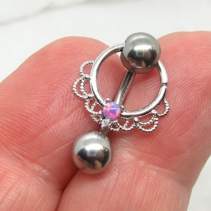 May include: A silver-colored body piercing jewelry piece. It features a circular design with a dangling ball and a small, pink opal-like stone. The jewelry is held up by a barbell and a decorative, scalloped edge.