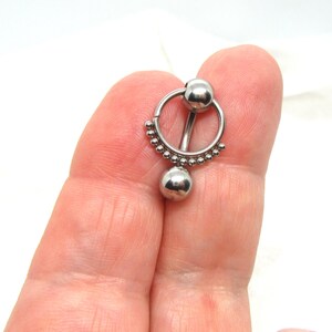 May include: A silver-colored body piercing jewelry piece. The jewelry features a circular design with a small ball at the top, a straight bar, and a larger ball at the bottom. The circular part is adorned with small, evenly spaced beads.