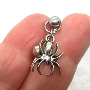 May include: A silver-colored spider charm with a small ball at the top, attached to a silver loop. The spider has eight legs and a rounded body. The charm is designed to be worn as jewelry.