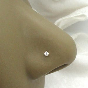 May include: A small, round, clear gemstone nose stud set in a metallic prong setting. The jewelry is positioned on the side of a nose, featuring a single faceted stone that reflects light, creating a bright, sparkling appearance on the skin.