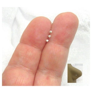 May include: Three small, clear, cubic zirconia nose rings on a finger. The rings are set in silver.