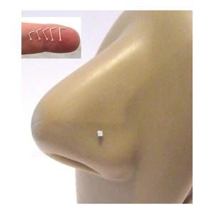 Puede incluir: Un piercing de nariz de plata con una pequeña piedra preciosa transparente de corte cuadrado en un engaste de garras. La joya se muestra insertada en una nariz de modelo, con una imagen insertada que muestra cinco piercings en L de plata adicionales.