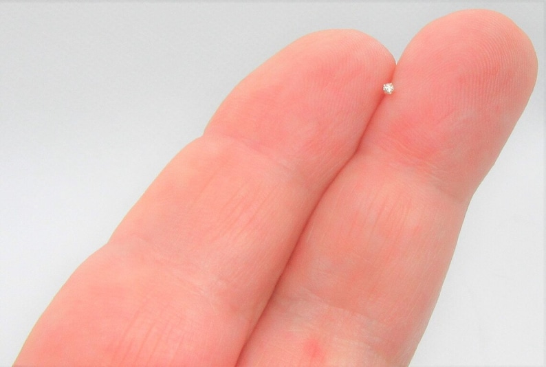 May include: A small, clear, round diamond gemstone held between two fingers.