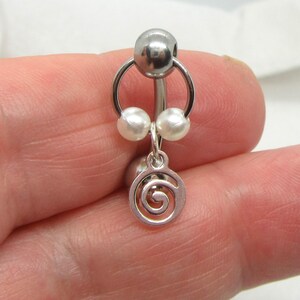 May include: A silver belly button ring with a spiral charm. The ring has a large top ball, two small pearl-like beads, and a dangling spiral design. The jewellery is designed for body piercing.