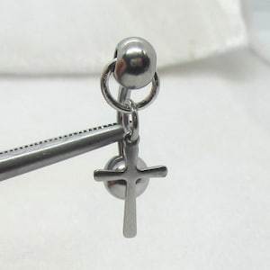 May include: A silver-colored, dangling cross earring. The earring features a small cross pendant suspended from a curved barbell with a ball on each end. The earring is held by a silver-colored tool.