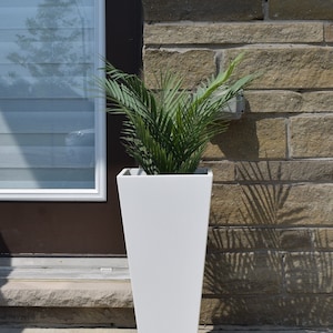 May include: A white, tall, tapered planter with a green plant inside. The planter is sitting on a concrete surface in front of a brick wall.