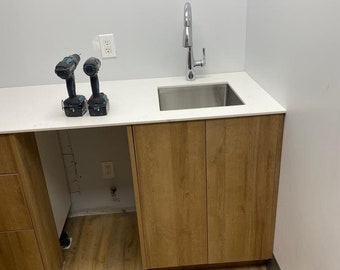 May include: A white countertop with a stainless steel sink and a chrome faucet. Two black and blue cordless drills are on the countertop. The countertop is attached to a set of brown cabinets.