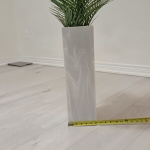 以下が含まれることがあります： 大理石模様の白いセラミックの花瓶に、緑の人工ヤシの木が植えられています。花瓶の横に、黄色のメジャーが置かれています。
