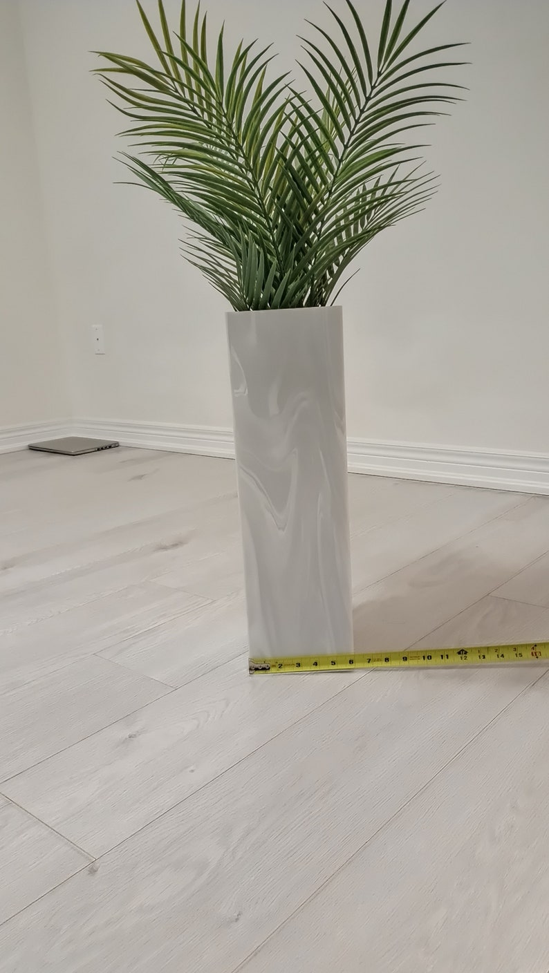 以下が含まれることがあります： 大理石模様の白いセラミックの花瓶に、緑の人工ヤシの木が植えられています。花瓶の横に、黄色のメジャーが置かれています。