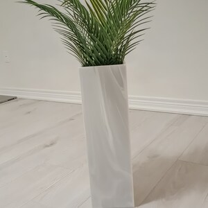 以下が含まれることがあります： 渦巻き模様の白いセラミックの花瓶に、緑の葉の植物が入っています。花瓶は白い木製の床の上に置かれています。