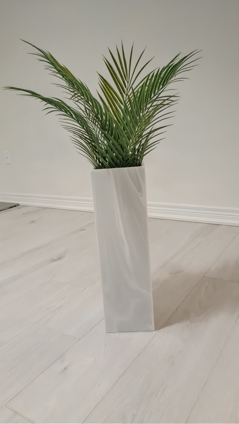 以下が含まれることがあります： 渦巻き模様の白いセラミックの花瓶に、緑の葉の植物が入っています。花瓶は白い木製の床の上に置かれています。