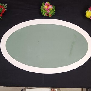 May include: An oval-shaped mirror with a white frame, set against a black backdrop. The mirror's surface is a muted green. Decorative floral arrangements in vases are placed around the mirror, adding pops of color.