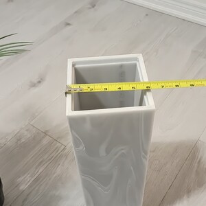 以下が含まれることがあります： 大理石模様の白いセラミックの花瓶。花瓶は正方形で、上部に長方形の開口部があります。黄色のメジャーが花瓶に巻き付けられており、15.2cmの測定値が表示されています。