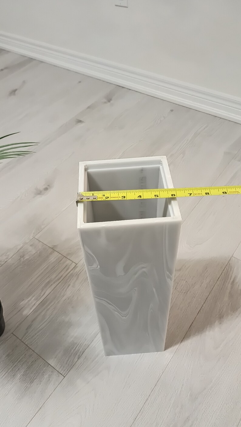 以下が含まれることがあります： 大理石模様の白いセラミックの花瓶。花瓶は正方形で、上部に長方形の開口部があります。黄色のメジャーが花瓶に巻き付けられており、15.2cmの測定値が表示されています。