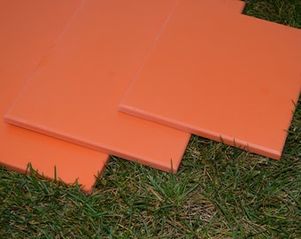 May include: Four orange square stepping stones are arranged on a bed of green grass. The stones are a solid, matte orange color and appear to be made of a durable material. The edges are slightly rounded.