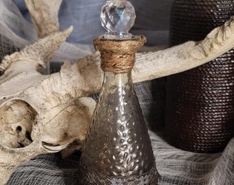 Puede incluir: Una botella de vidrio decorativa con una superficie texturizada y un tapón de cristal. La botella está adornada con cuerda y una banda decorativa. Una segunda botella y una calavera están en el fondo, lo que sugiere un tema gótico o de boticario.