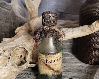 Pode incluir: Uma garrafa de vidro verde escuro rotulada "Monkshood" com uma tampa decorativa e barbante. A garrafa é adornada com um rótulo com ilustrações botânicas. O fundo inclui um crânio e tecido texturizado.