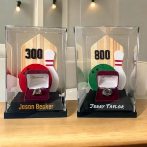 May include: Two clear acrylic display cases with black bases. Each case contains a bowling pin, bowling ball, and a ring in a red velvet box. The first case has a score of 300 and the name "Jason Becker". The second case has a score of 800 and the name "Jerry Taylor".