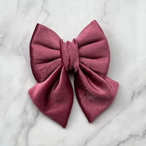 Puede incluir: Un lazo de terciopelo burdeos, sobre una superficie de mármol. El lazo es de un tono profundo y rico de rojo-púrpura, con un aspecto suave y texturizado. El diseño del lazo presenta un nudo central y dos bucles simétricos.