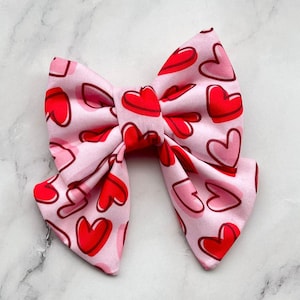 Può includere: Un fiocco di stoffa rosa con un motivo a cuori rossi. Il fiocco è legato in una forma classica con due anelli e un nodo centrale.