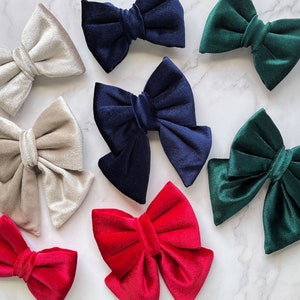 May include: Assortment of velvet hair bows in various colours, including navy blue, green, beige, and red. The bows are arranged on a white marble surface.