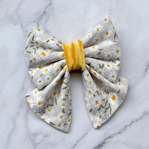 Può includere: Un fiocco per capelli in tessuto con un motivo floreale a margherita bianco e giallo su sfondo grigio chiaro. Il centro del fiocco è una fascia gialla brillante e strutturata. Questo accessorio è adatto a varie occasioni.
