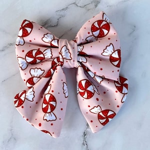 May include: A large, pink hair bow with a repeating pattern of red and white peppermint candies. The bow is tied in the center and has two large loops on each side. Small red dots are scattered across the bow.