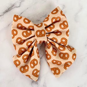 Può includere: Un grande fiocco di colore crema con un motivo di pretzel marroni. Il fiocco è fatto di tessuto ed è legato in una classica forma di fiocco.