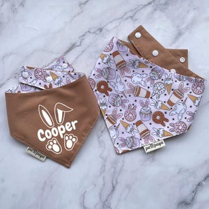 Könnte beinhalten: Zwei braune und weiße Bandana-Lätzchen mit einem rosa und weißen Muster. Ein Lätzchen trägt den Namen "Cooper" und ein Hasen-Design. Das andere Lätzchen hat ein Muster aus Donuts, Eiswaffeln und Cupcakes.
