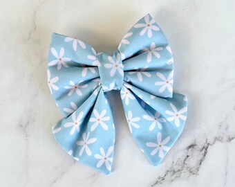 May include: A light blue bow with a white daisy print. The bow is made of fabric and has a simple, elegant design.