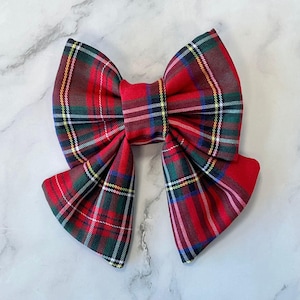 May include: A large, red plaid bow with green, blue, and yellow accents. The bow is made of fabric and has a classic shape, suitable for hair accessories or gift wrapping.