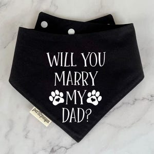 Op de afbeelding: Zwarte bandana met witte poot afdrukken en de tekst "Wil je met mijn papa trouwen?"