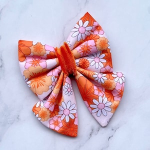 Puede incluir: Un gran lazo floral naranja y blanco con un centro de terciopelo. El lazo está hecho de tela con un estampado de margaritas.