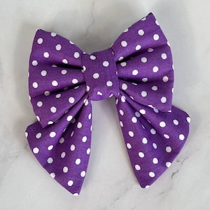 Può includere: Un fiocco viola con pois bianchi. Il fiocco è fatto di tessuto ed è legato in una classica forma di fiocco.