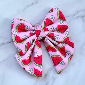 Può includere: Un fiocco di stoffa rosa con stampa di anguria e fiori bianchi. Il fiocco è realizzato in tessuto morbido ed è perfetto per aggiungere un tocco di divertimento estivo a qualsiasi outfit.