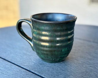 May include: Handmade ceramic mug with a dark green and black glaze. The mug has a textured, ridged design and a curved handle. The interior is a darker shade of black. A unique piece for enjoying coffee or tea.