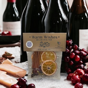 May include: A close-up of a small brown paper bag with the text "Warm Wishes - Mulling Spice Pack" printed on it. The bag contains dried orange slices, cinnamon sticks, and other spices. The bag is sitting on a table with a bottle of red wine, a glass of red wine, and a bowl of cranberries.