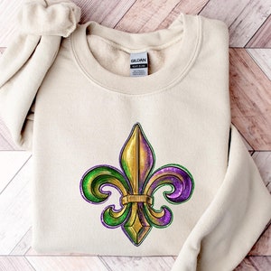 Puede incluir: Una sudadera beige con un diseño de flor de lis en oro, verde y morado. La sudadera está doblada y colocada sobre una superficie de madera.