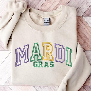 May include: A beige sweatshirt with the words "Mardi Gras" in green, yellow, and purple letters.