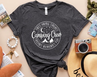 May include: A gray t-shirt with a white circle design that says "Camping Crew" with a campfire, trees, and stars. The shirt is laid out on a beige surface with other travel items, including a straw bag, a passport, a map, sunglasses, a camera, and a straw hat with the text "bon voyage" on it.