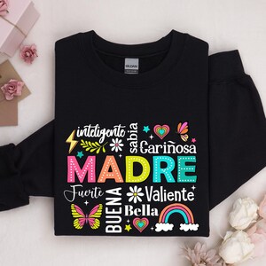 May include: Black sweatshirt with a colorful graphic design that says "MADRE" surrounded by words in Spanish, including "inteligente", "sabia", "cari&ntilde;osa", "fuerte", "buena", "bella", and "valiente".