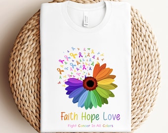Op de afbeelding: Wit T-shirt met een kleurrijk bloemendesign gemaakt van linten. De bloem heeft een zwart midden en de linten zijn in verschillende kleuren, waaronder roze, blauw, groen, geel en oranje. De tekst "Faith Hope Love Fight Cancer In All Colors" is onder de bloem gedrukt.