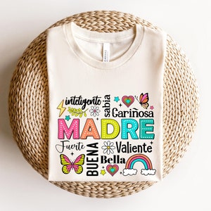 May include: A white t-shirt with a colorful graphic design that says "inteligente, sabia, cari&ntilde;osa, MADRE, fuerte, buena, bella, valiente". The graphic includes butterflies, hearts, stars, and a rainbow.