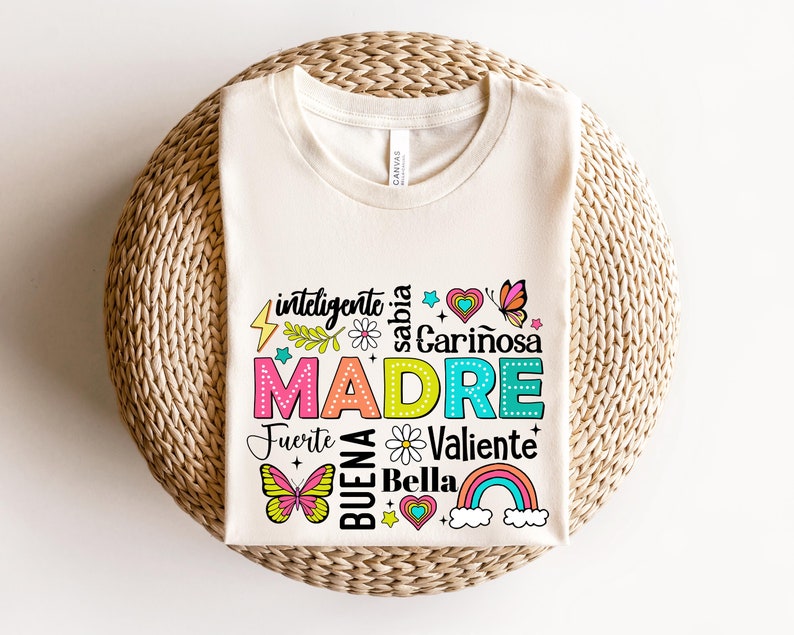 May include: A white t-shirt with a colorful graphic design that says "inteligente, sabia, cari&ntilde;osa, MADRE, fuerte, buena, bella, valiente". The graphic includes butterflies, hearts, stars, and a rainbow.
