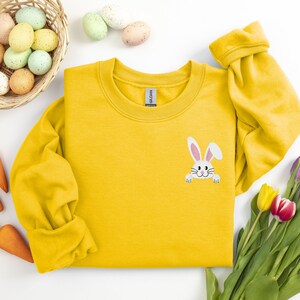 May include: A yellow sweatshirt with a white bunny peeking out from the pocket. The bunny has pink ears and nose. The sweatshirt is surrounded by Easter eggs, carrots, and tulips.
