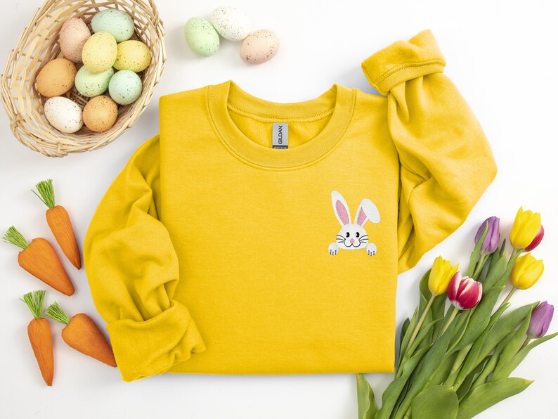 May include: A yellow sweatshirt with a white bunny peeking out from the pocket. The bunny has pink ears and nose. The sweatshirt is surrounded by Easter eggs, carrots, and tulips.