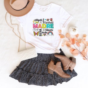 May include: White t-shirt with a colorful graphic design that says "MADRE" surrounded by Spanish words describing a mother, including "inteligente", "sabia", "cari&ntilde;osa", "fuerte", "buena", "bella", and "valiente".