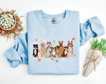 May include: Light blue sweatshirt with a graphic of a group of dogs wearing bunny ears and holding Easter eggs. The dogs are surrounded by flowers and a carrot.